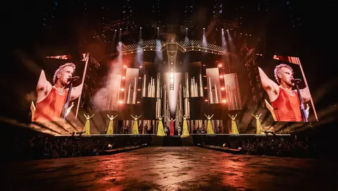 A performer in a red outfit sings on stage, flanked by dancers, with large screens displaying visuals and dramatic lighting effects.