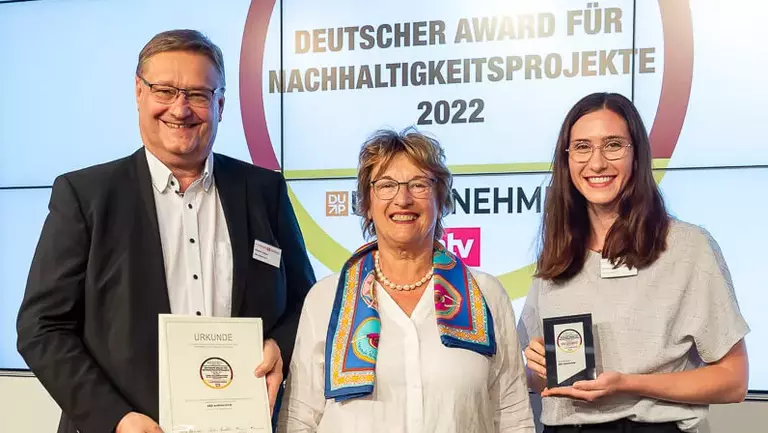 Drei Personen stehen zusammen und halten Auszeichnungen bei der Veranstaltung Deutscher Award für Nachhaltigkeitsprojekte 2022, vor einem Hintergrund, der das Logo der Veranstaltung zeigt.