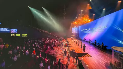 A large crowd gathers in a concert hall, with performers on stage and colorful lighting illuminating the scene. Text is displayed on a large screen.