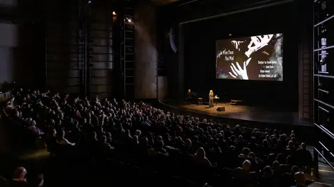 A speaker presents on stage in front of a large audience, with a screen displaying text and visuals related to the topic.