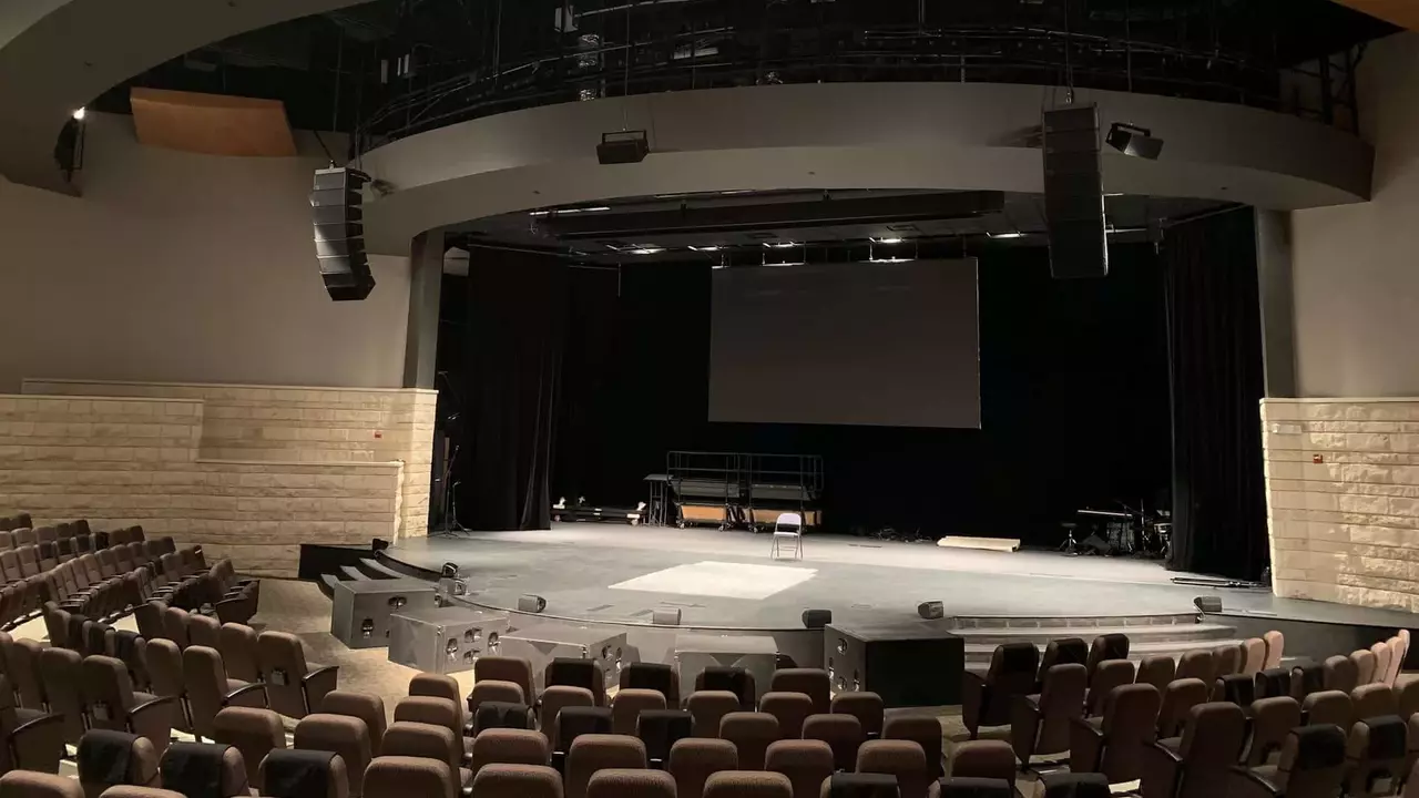 Empty theater stage with seating arranged in a semi-circle, featuring a large screen and audio equipment overhead.