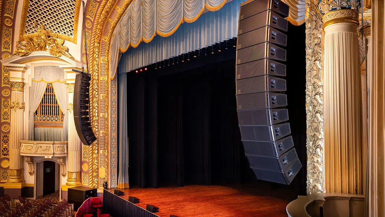 Ornate theater interior featuring a wooden stage, red seating, and large sound speakers suspended above the stage.