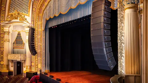 Ornate theater interior featuring a wooden stage, red seating, and large sound speakers suspended above the stage.