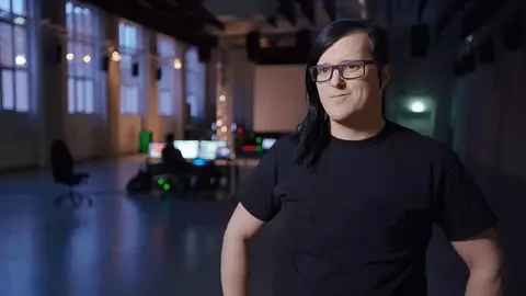 A person with long dark hair and glasses stands in a dimly lit room, with computer equipment visible in the background.