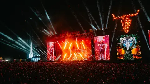 Large crowd at a music festival with bright stage lights and visual displays, featuring a prominent bull sculpture above the stage.