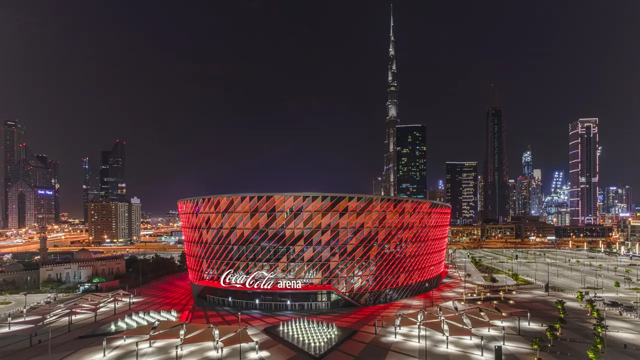 Coca-Cola Arena nachts beleuchtet, mit modernem Design und roten Akzenten, umgeben von Dubais Skyline und Wolkenkratzern.