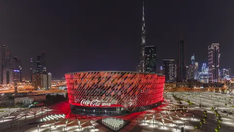 Coca-Cola Arena illuminated at night, featuring a modern design with red accents, surrounded by Dubai's skyline and skyscrapers.