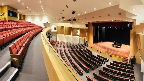 Interior of a theater featuring red seating, a stage with a grand piano, and a well-lit auditorium layout.
