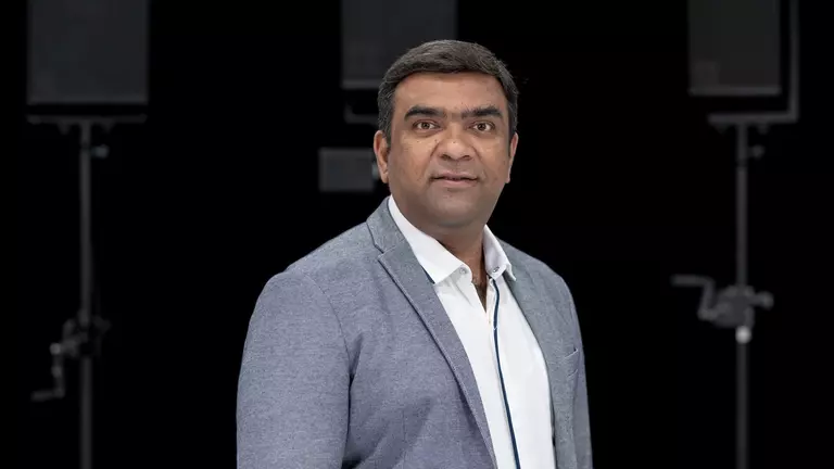 Man in a gray blazer stands confidently, facing the camera, with a neutral background featuring audio equipment.