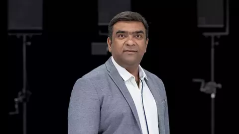 Man in a gray blazer stands confidently, facing the camera, with a neutral background featuring audio equipment.