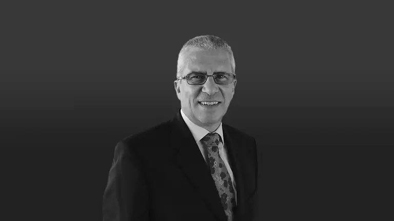 Man in a suit with glasses, smiling against a dark background. He has short, light-colored hair and is wearing a patterned tie.