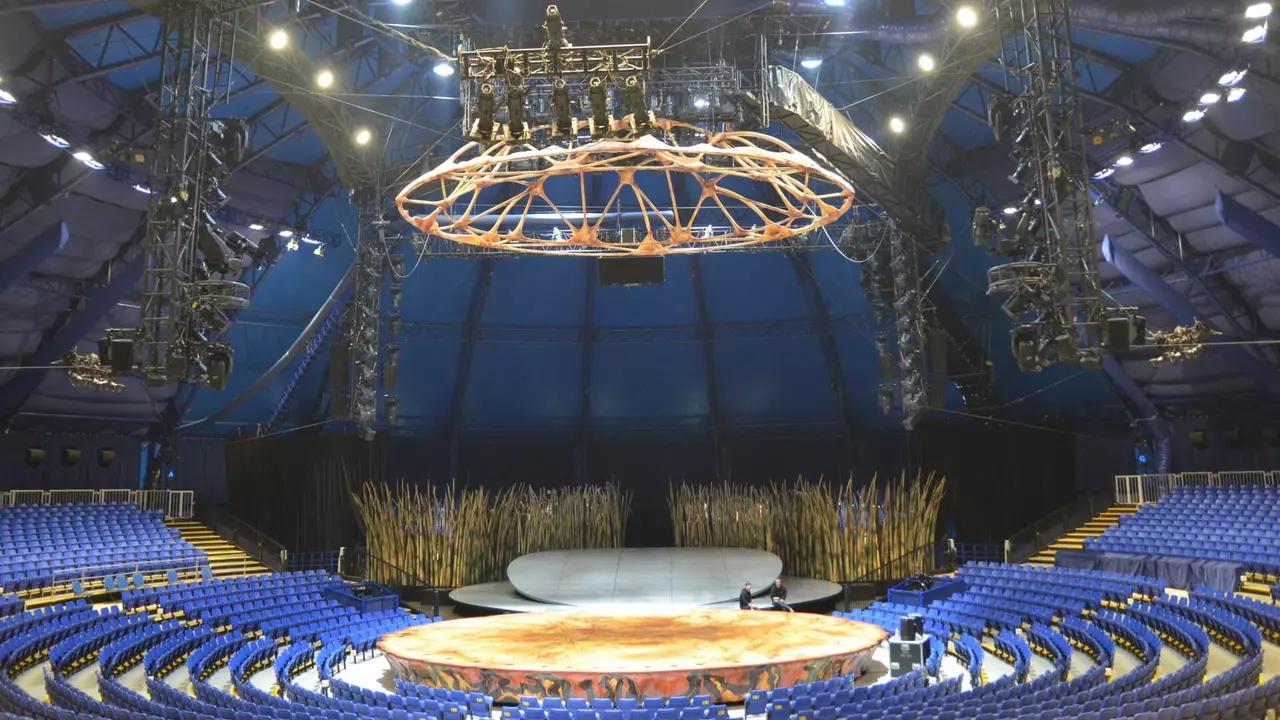 Circus tent interior featuring a circular stage at the center, surrounded by blue seating and overhead lighting fixtures.
