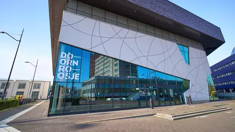 Modern building with large glass windows reflecting surrounding structures, featuring a geometric design and the text "DOORNROOSJE" prominently displayed.