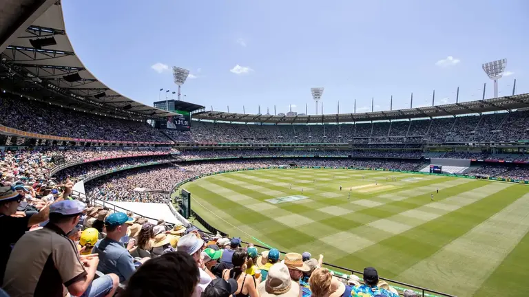 Crowd gathered in a stadium watching a cricket match, with players visible on the field and bright blue skies overhead.