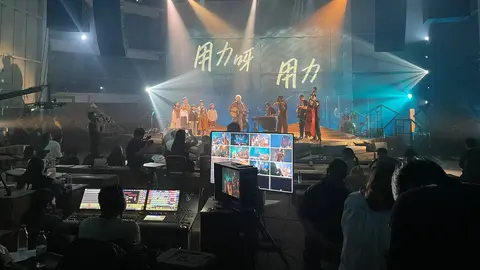 A live performance on stage with musicians and dancers, illuminated by colorful lights, while a control panel displays video feeds.