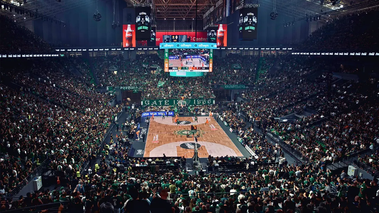 A packed basketball arena filled with fans, featuring a court in the center and a large scoreboard displaying game information.