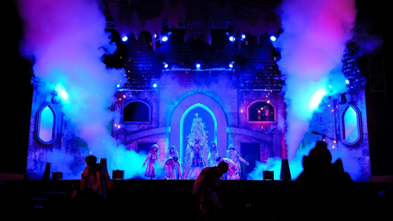 A theatrical performance on stage with dancers in colorful costumes, illuminated by blue and purple lights, surrounded by smoke effects.