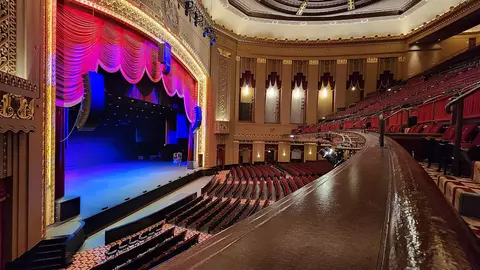 Interior view of a theater featuring a stage with red curtains and blue lighting, surrounded by rows of empty seats.