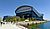 Gugon Arena in Korea, a modern architectural structure, surrounded by greenery and a waterfront promenade, with people walking nearby.