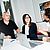 Three professionals engage in a discussion at a conference table, with laptops and a presentation screen in the background.