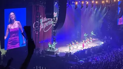 A performer in a pink dress dances on stage, accompanied by a group of dancers, with a large screen displaying her image in the background.