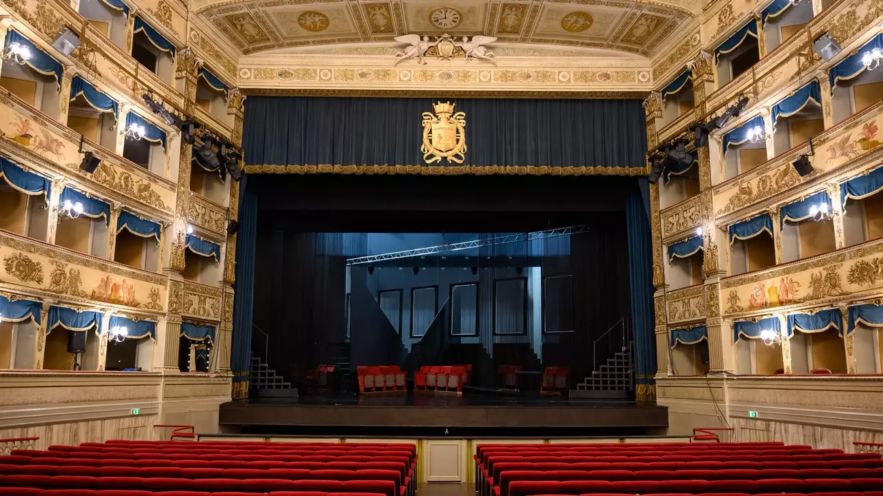 Interior of a theater featuring ornate balconies, red seating, and a dark stage with a blue curtain and decorative elements.