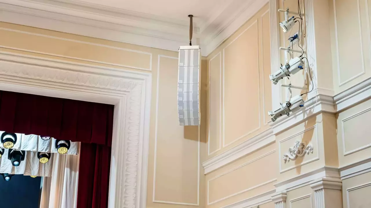 Ceiling-mounted sound system speaker installed on a decorative wall in a theater, with stage curtains visible in the background.