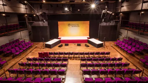 Theater stage with a central podium, surrounded by rows of purple chairs, illuminated by stage lights.