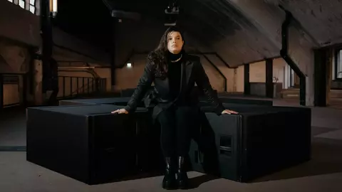 A woman sits on large speaker boxes in an industrial space, wearing a black outfit and looking directly at the camera.