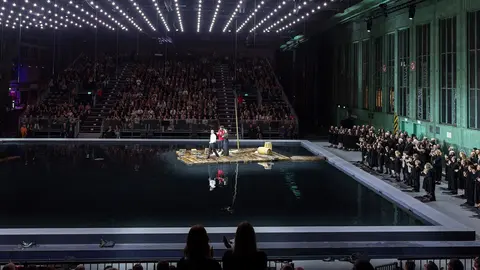 A large audience watches a performance on a stage with a water feature, illuminated by overhead lights in an industrial setting.