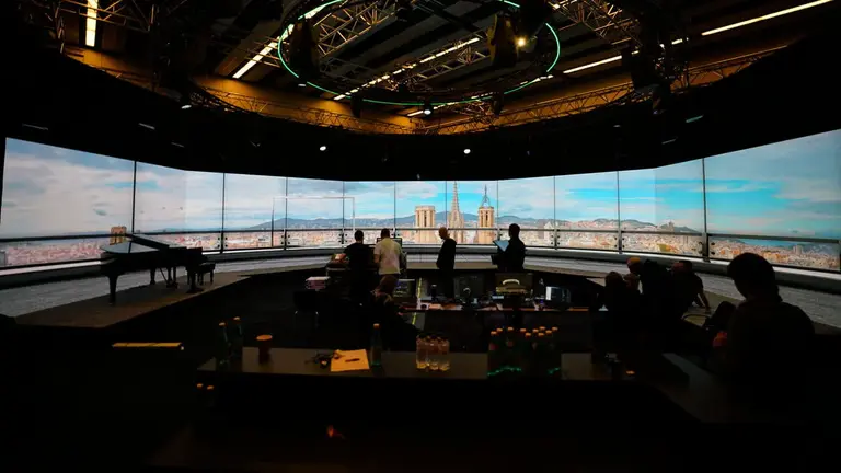 Modern studio interior with a panoramic screen displaying a cityscape, featuring people engaged in various activities and a piano in the foreground.