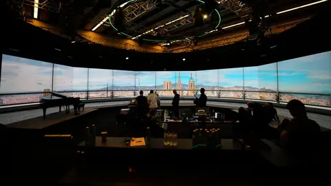 Modern studio interior with a panoramic screen displaying a cityscape, featuring people engaged in various activities and a piano in the foreground.