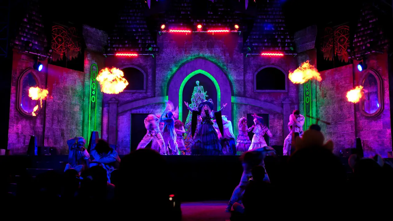 A theatrical performance featuring performers in elaborate costumes on stage, with colorful lighting and fire effects in the background.