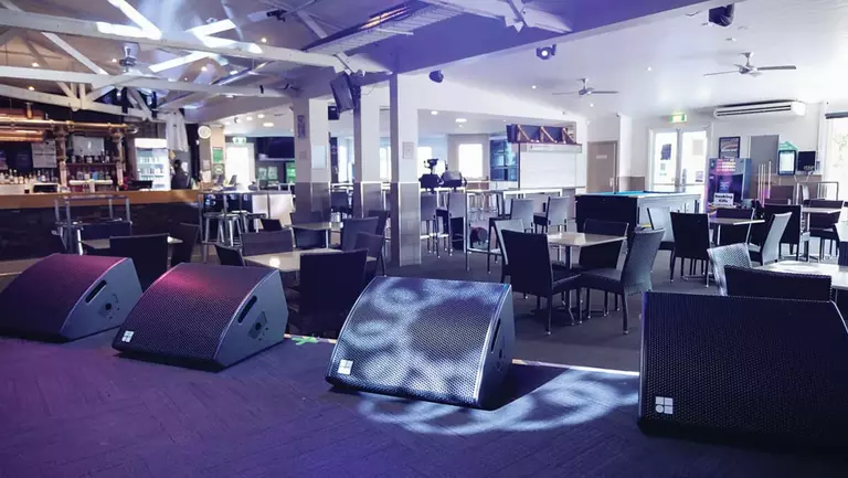 Empty venue with sound speakers in the foreground, tables and chairs arranged for an event, and a bar area visible in the background.
