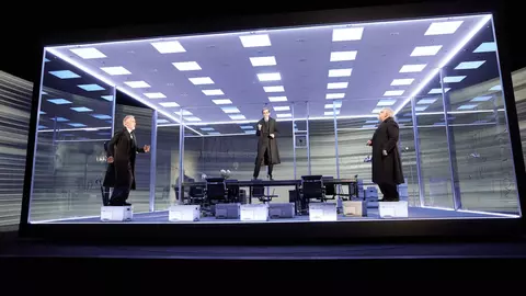 Three figures in formal attire stand in a modern, illuminated glass office space, with one figure elevated above the others.