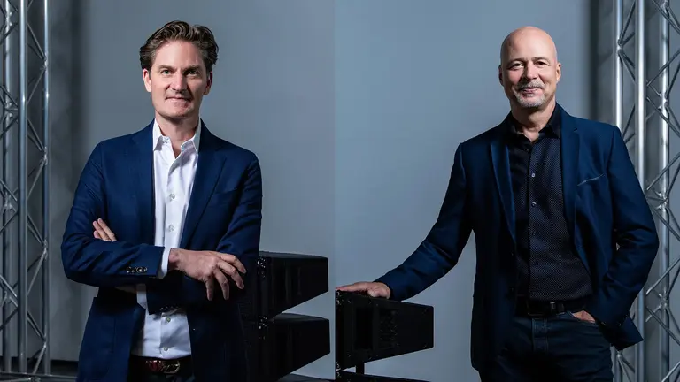 Zwei Männer in Businesskleidung stehen nebeneinander, jeder mit einer selbstbewussten Haltung, vor einem neutralen Hintergrund mit geometrischen Strukturen.