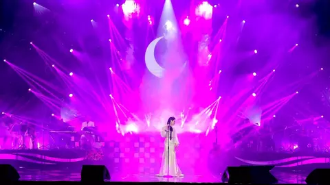 A performer stands on stage in a flowing white outfit, surrounded by vibrant purple lights and a crescent moon backdrop.