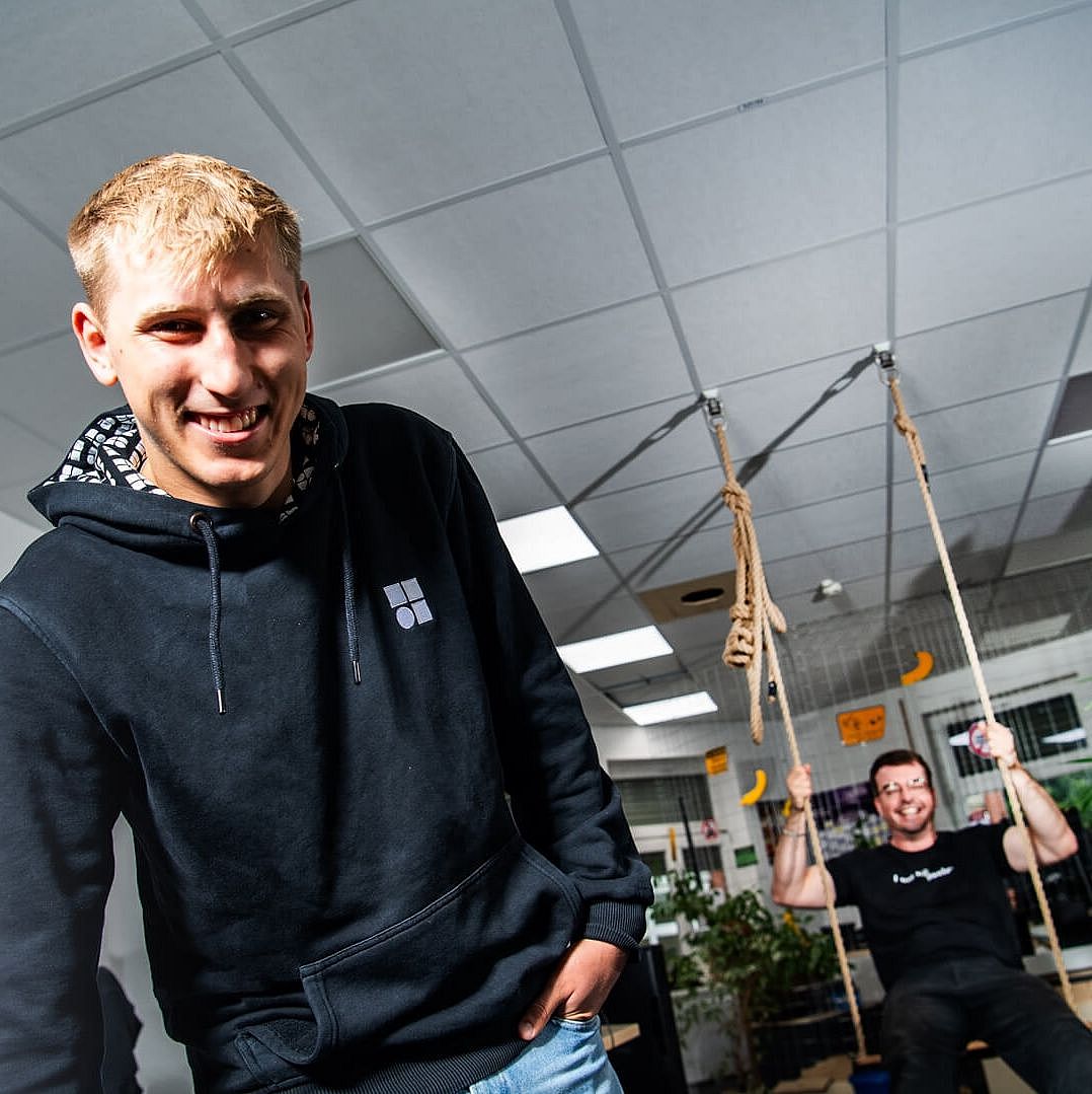 Two young men in an office setting; one stands smiling in a hoodie, while the other swings on a rope swing in the background.