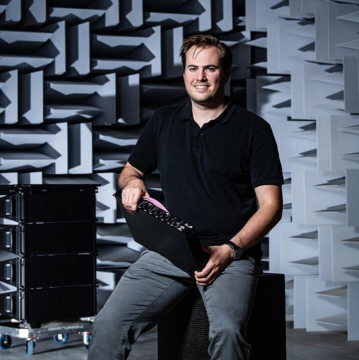 A man sits on a speaker in a soundproof room, holding a device with a pink handle, against a textured wall.
