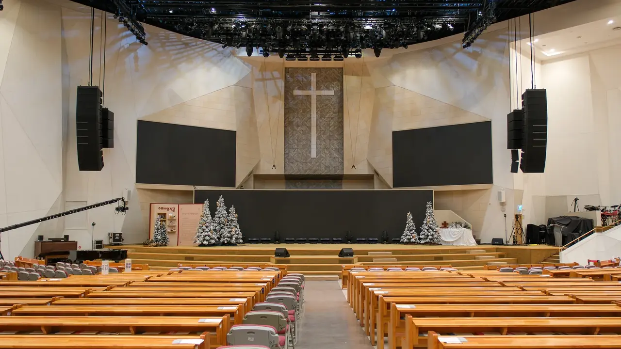 Interior of a church auditorium featuring wooden pews, a large cross on the wall, and two blank screens flanking the stage.