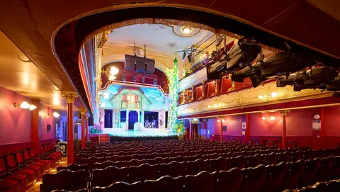 Interior of a theater featuring rows of empty seats facing a brightly lit stage decorated for a performance, with colorful lights and a festive backdrop.