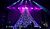A performer stands atop a large, patterned structure on stage, surrounded by musicians and illuminated by purple and blue lights.