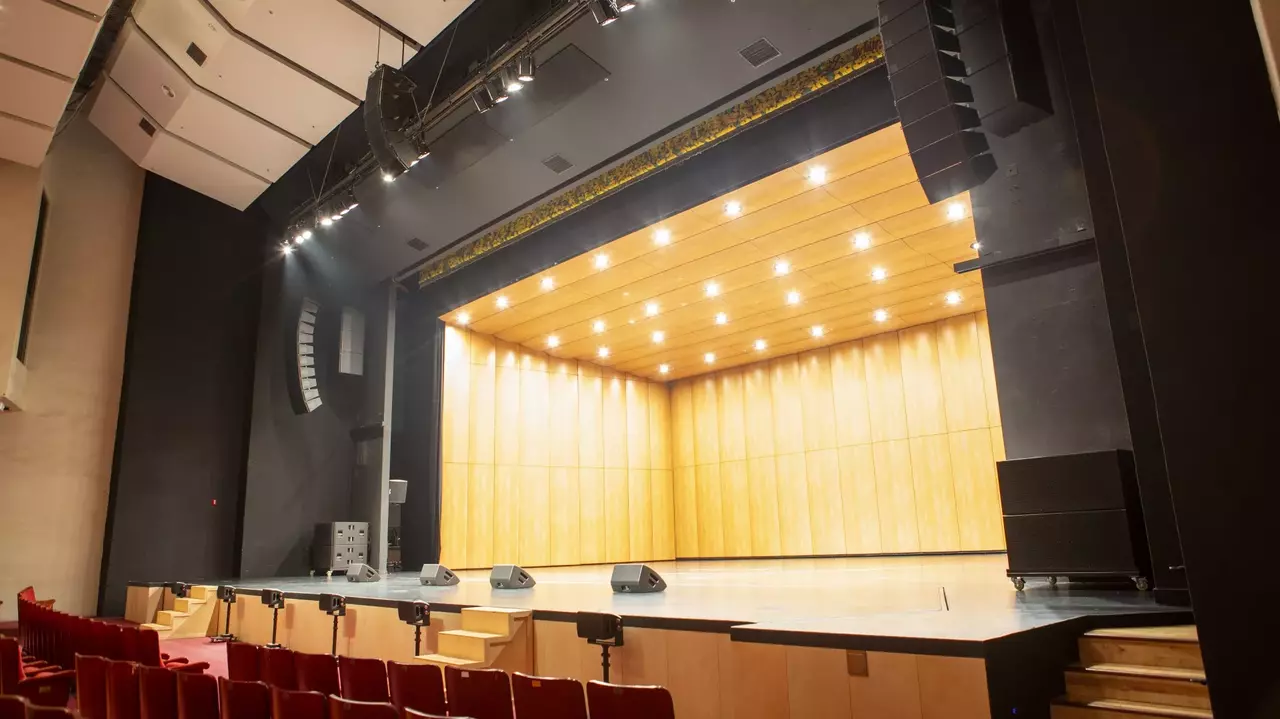Empty auditorium stage with wooden paneling and overhead lights, featuring sound equipment and red seating in the foreground.