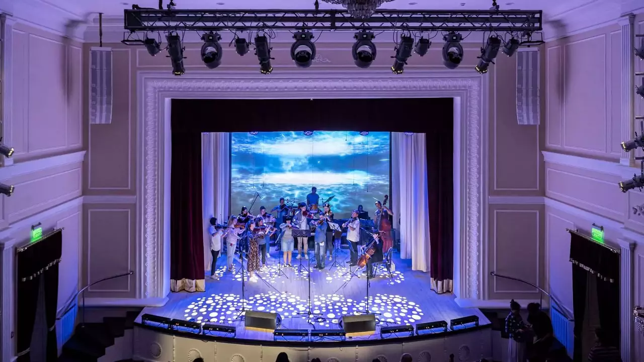 A musical ensemble performs on stage in a theater, with a large screen displaying a scenic background and an audience seated in front.