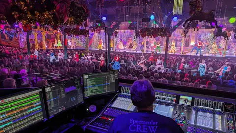 A sound technician operates mixing equipment while a colorful stage performance takes place in front of a large audience.