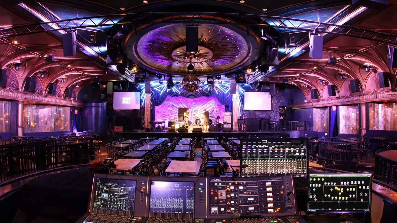A concert venue featuring a stage with lighting, sound equipment in the foreground, and rows of tables arranged for an audience.
