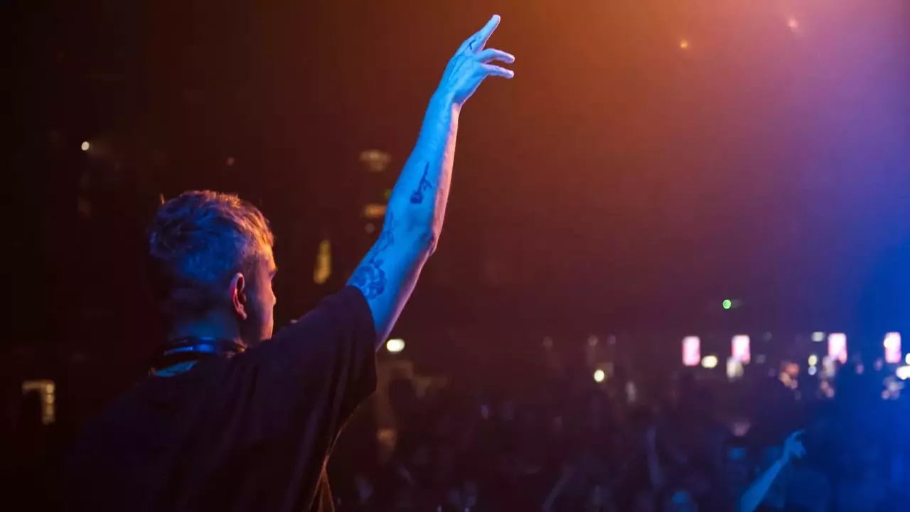 DJ, der auf der Bühne auftritt, mit einer Hand erhoben, umgeben von einem Publikum in einem schwach beleuchteten Veranstaltungsort.