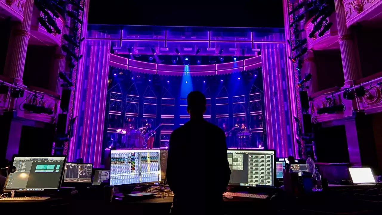 A silhouette of a person stands in front of a control desk, overlooking a brightly lit stage with purple lighting and a band setup.