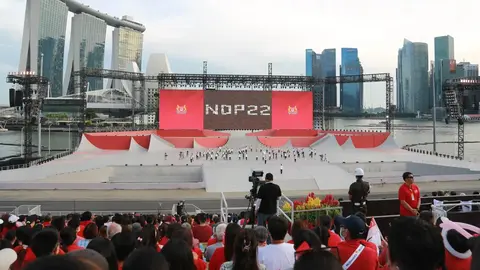 A large outdoor stage set up for the National Day Parade 2022, with a crowd in red attire and a skyline of modern buildings in the background.
