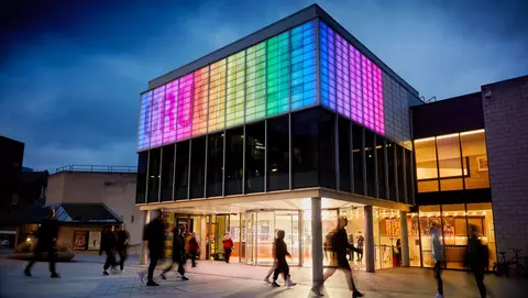 Modern building with colorful illuminated facade, people walking in front during twilight.